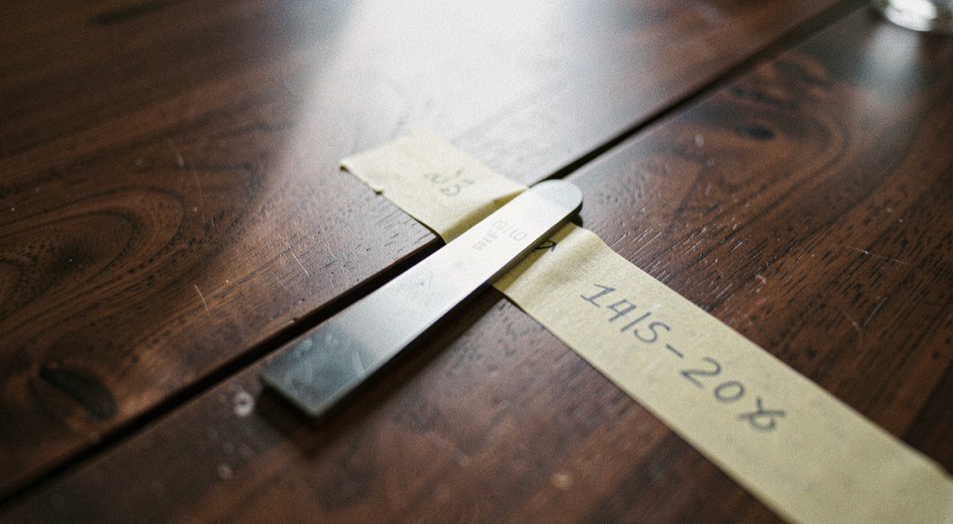 A steel feeler gauge blade slipped into a long split in a dark walnut table top with a strip of masking tape beside it carrying a pencilled measurement, lamp light raking across the surface, close view.
