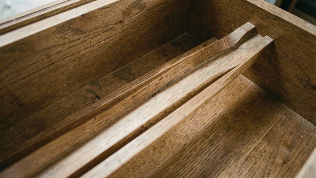The inside of an oak chest of drawers carcase with a timber runner rail worn into a deep groove along its middle, a torch beam picking out the hollow, close view into the carcase.